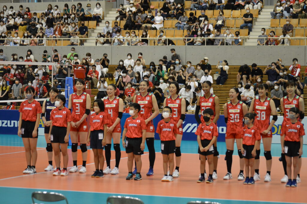 公式戦ユニフォームに着替え笑顔の女子バレー日本代表選手達とその前に立つ子ども達